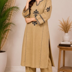 May include: A tan-coloured long-sleeved tunic and matching wide-leg trousers set. The tunic features a notched neckline, black embroidered floral motifs on the chest, and black geometric trim along the cuffs, hem, and vertical front panels.