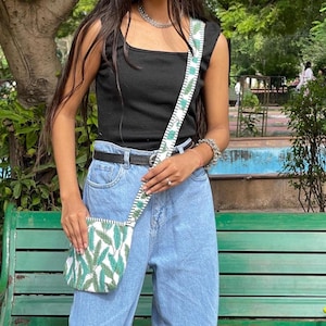 Puede incluir: Un pequeño bolso cruzado blanco y verde con un estampado de hojas. El bolso tiene una correa a juego y se lleva al hombro. La persona lleva un top negro y vaqueros azules.