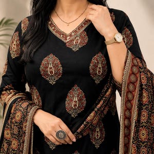 May include: A black long-sleeved tunic featuring a V-neckline with a decorative border and a repeating teardrop-shaped floral pattern in shades of red, cream, and gold. The garment is paired with a matching shawl that has a dense floral border.