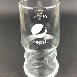 Könnte beinhalten: Ein klares Glas Pepsi-Trinkglas mit einem weißen Pepsi-Logo und dem Wort "Pepsi" auf dem Boden. Das Glas hat ein einzigartiges, gedrehtes Design.