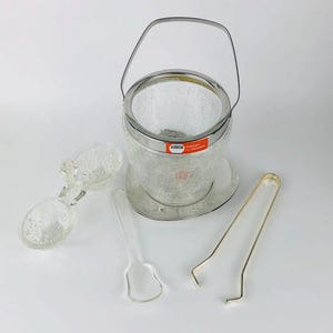May include: A clear glass ice bucket with a silver handle and a matching base. The bucket has a textured surface and comes with two clear glass spoons and a pair of gold tongs.