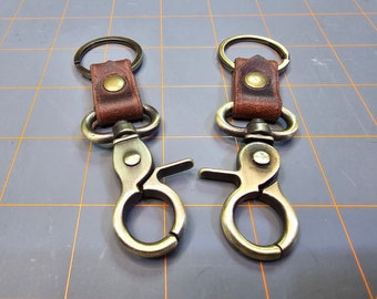 May include: Two antique brass keychains with brown leather straps and metal rings. The keychains have a swivel clasp.