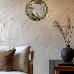 May include: A round, ornate gold mirror hangs on a textured wall. The mirror reflects a glimpse of a person. A black vase with dried plants sits on a dark surface. A light-colored sofa with brown and white pillows is in the foreground.