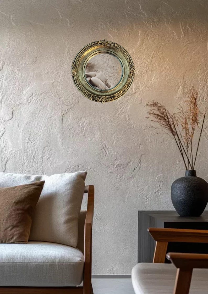 May include: A round, ornate gold mirror hangs on a textured wall. The mirror reflects a glimpse of a person. A black vase with dried plants sits on a dark surface. A light-colored sofa with brown and white pillows is in the foreground.