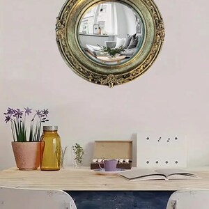 May include: A round, antique-style mirror with an ornate, gold-colored frame. The mirror reflects a living room scene. The image also shows a desk with a plant, a jar, and other decorative items.