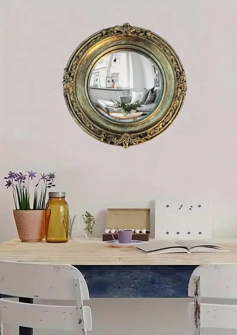 May include: A round, antique-style mirror with an ornate, gold-colored frame. The mirror reflects a living room scene. The image also shows a desk with a plant, a jar, and other decorative items.