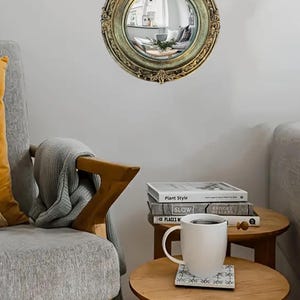 May include: A round, antique-style mirror with a gold-colored ornate frame. The mirror reflects a room with furniture. The mirror is hanging on a white wall. The room has a cozy, vintage aesthetic.