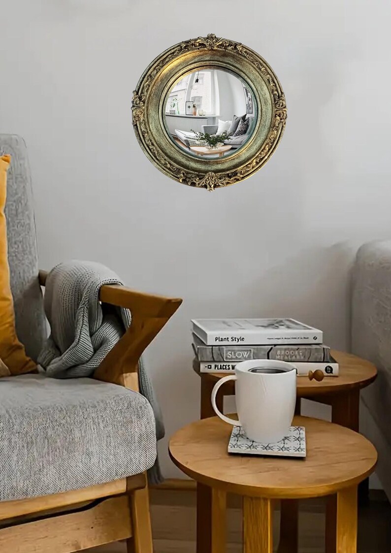 May include: A round, antique-style mirror with a gold-colored ornate frame. The mirror reflects a room with furniture. The mirror is hanging on a white wall. The room has a cozy, vintage aesthetic.