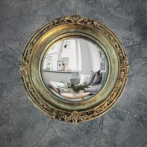 May include: A round, ornate mirror with a gold-colored frame. The mirror reflects a room with a couch, pillows, and a coffee table. The mirror is mounted on a textured gray wall.