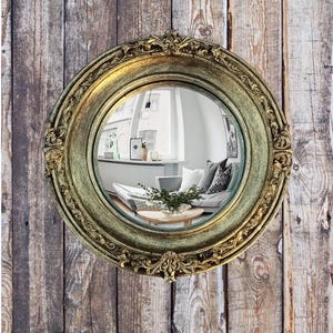 May include: A round, antique-style mirror with a gold-colored, ornate frame. The mirror reflects a room with a sofa, pillows, and a coffee table. The mirror is mounted on a wooden plank wall.