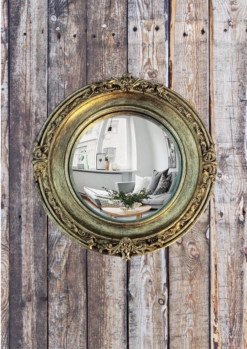 May include: A round, antique-style mirror with a gold-colored, ornate frame. The mirror reflects a room with a sofa, pillows, and a coffee table. The mirror is mounted on a wooden plank wall.