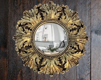 May include: A round, ornate gold mirror with a detailed leaf design around the edge. The mirror reflects a room with white walls, a couch, and a window. The mirror is mounted on a dark brown wooden wall.