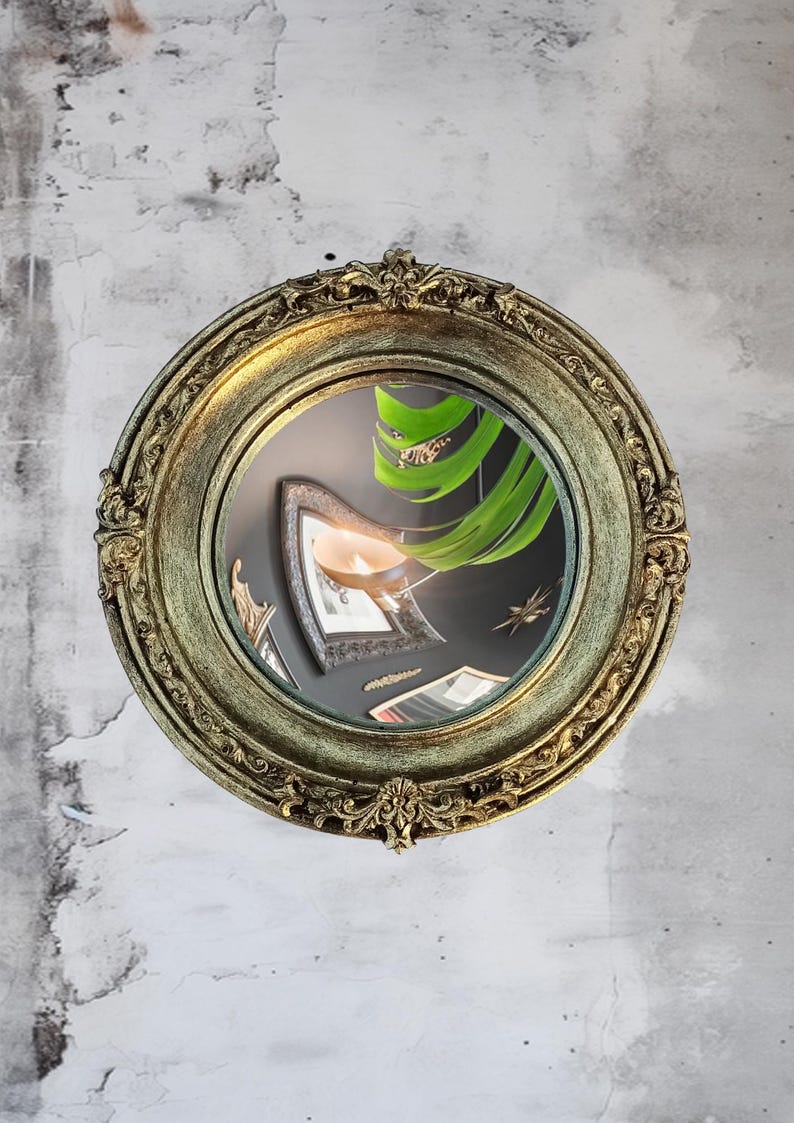 May include: A round, antique-style mirror with a gold-colored, ornate frame. The mirror reflects a dark interior with other framed objects and a large green plant. The mirror is mounted on a textured, light gray wall.