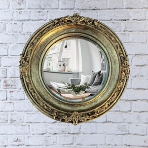 May include: A round, antique-style mirror with a gold-colored, ornate frame. The mirror reflects a room with a couch, coffee table, and window. The mirror is mounted on a white brick wall.