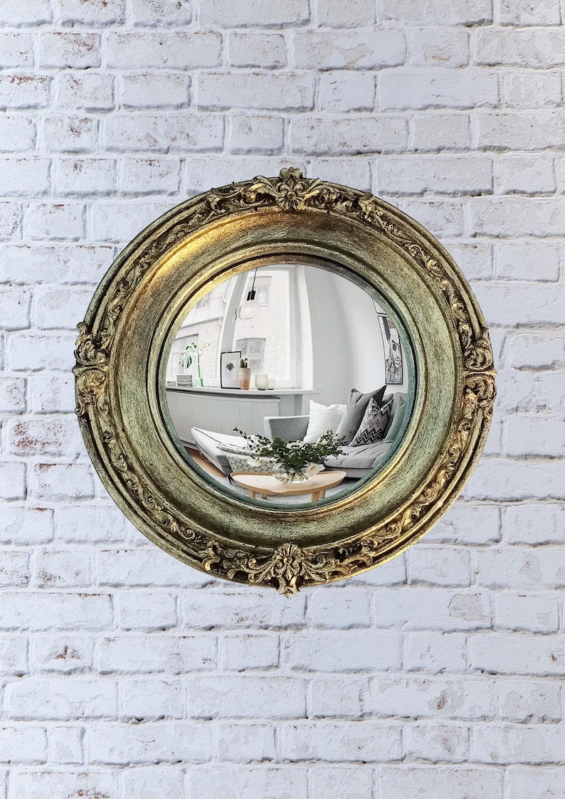 May include: A round, antique-style mirror with a gold-colored, ornate frame. The mirror reflects a room with a couch, coffee table, and window. The mirror is mounted on a white brick wall.