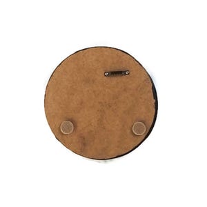 May include: A round, brown wooden plaque with a black edge. The back of the plaque features a metal hanger and two small, round, tan-colored pads. The plaque is set against a white background.