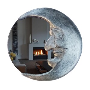 May include: A silver-colored crescent moon mirror with a textured surface. The mirror's reflective surface shows a room with a fireplace and furniture. The moon has a smiling face design.