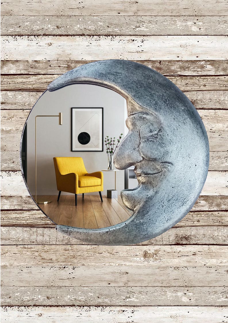 May include: A decorative wall mirror shaped like a crescent moon with a smiling face. The mirror has a silver finish and reflects a room with a yellow armchair, artwork, and a floor lamp. The background is a wood-plank wall.