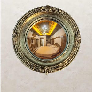 May include: A round, ornate mirror with a gold and green finish. The mirror reflects a bedroom with a bed, wooden furniture, and a ceiling with warm lighting. The mirror is mounted on a textured, off-white wall.