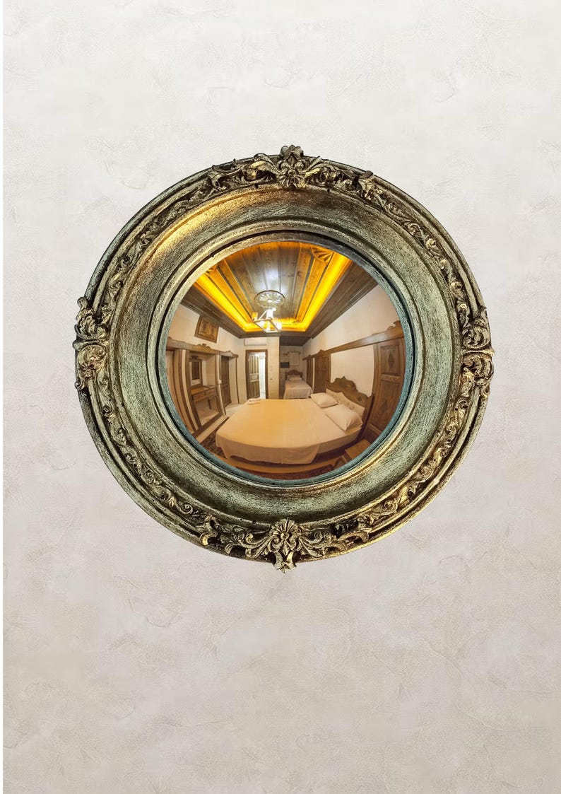 May include: A round, ornate mirror with a gold and green finish. The mirror reflects a bedroom with a bed, wooden furniture, and a ceiling with warm lighting. The mirror is mounted on a textured, off-white wall.