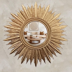 May include: A gold-colored sunburst mirror with a round central mirror reflecting a living room scene. The frame has a textured, radiating design. The mirror is mounted on a textured, light-colored wall.