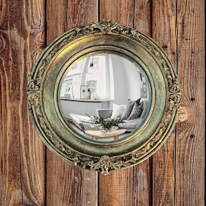 May include: A round, antique-style mirror with an ornate, gold-colored frame. The mirror reflects a living room scene with a sofa, pillows, and a coffee table. The mirror is mounted on a wooden wall with vertical planks.