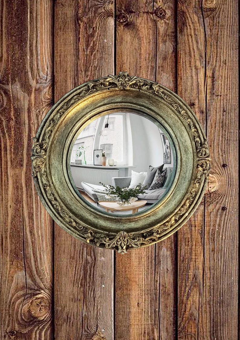 May include: A round, antique-style mirror with an ornate, gold-colored frame. The mirror reflects a living room scene with a sofa, pillows, and a coffee table. The mirror is mounted on a wooden wall with vertical planks.