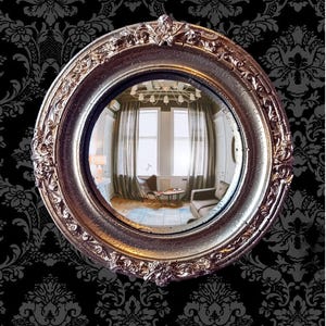 Può includere: Uno specchio convesso rotondo caratterizzato da una cornice metallica color oro ornata con intricati dettagli in rilievo. La superficie riflettente mostra una vista grandangolare distorta dell'interno di una stanza con finestre, tende e mobili.