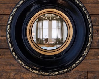May include: A round black mirror with a gold inner frame and decorative gold trim. The mirror reflects a room with windows, curtains, and furniture. The mirror is mounted on a wooden wall.