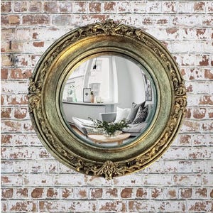 May include: A round, antique-style mirror with a gold-colored, ornate frame. The mirror reflects a room with a sofa, pillows, and a coffee table. The mirror is mounted on a brick wall.