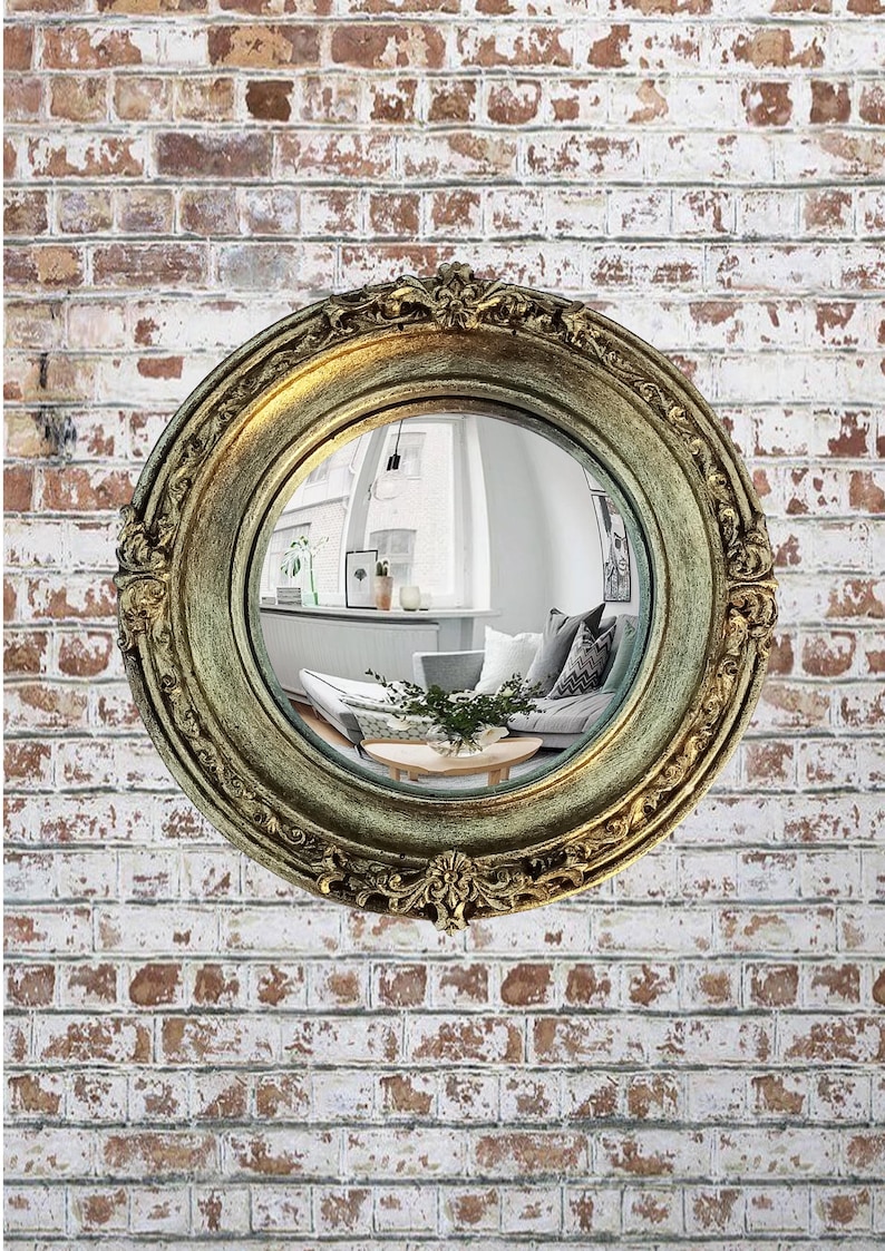 May include: A round, antique-style mirror with a gold-colored, ornate frame. The mirror reflects a room with a sofa, pillows, and a coffee table. The mirror is mounted on a brick wall.