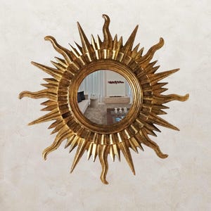 Può includere: Uno specchio a forma di sole dorato con uno specchio centrale rotondo e raggi radianti. Lo specchio ha un design decorativo e ornato ed è montato su una parete strutturata bianco sporco. Il design dello specchio ricorda un sole.
