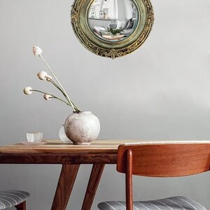 May include: A round, antique-style mirror with an ornate, gold-colored frame. The mirror reflects a room with furniture and decor. The mirror is hanging on a gray wall. A wooden table and chairs are in the foreground.