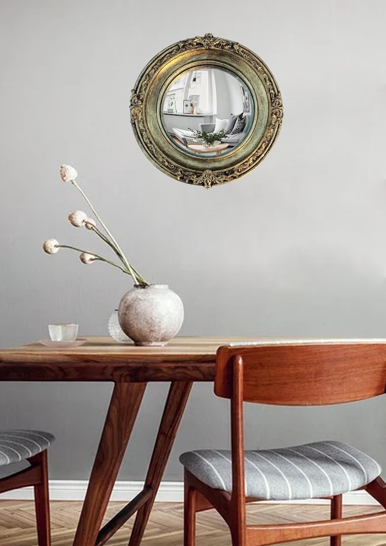 May include: A round, antique-style mirror with an ornate, gold-colored frame. The mirror reflects a room with furniture and decor. The mirror is hanging on a gray wall. A wooden table and chairs are in the foreground.