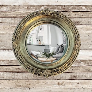 May include: A round, ornate mirror with a gold and green distressed finish. The mirror reflects a living room scene with a sofa, coffee table, and artwork. The mirror is mounted on a weathered, white-washed wood wall.