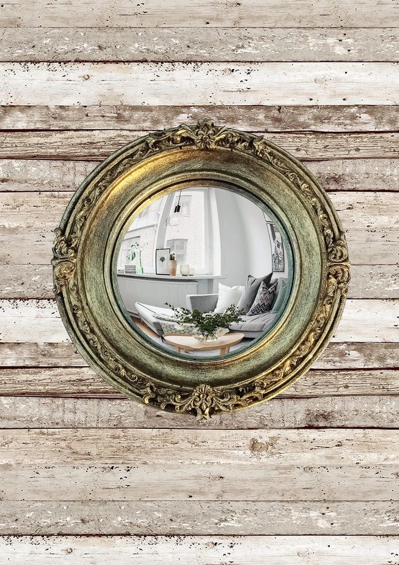 May include: A round, ornate mirror with a gold and green distressed finish. The mirror reflects a living room scene with a sofa, coffee table, and artwork. The mirror is mounted on a weathered, white-washed wood wall.