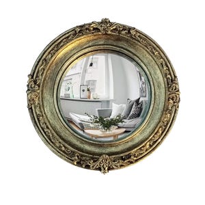May include: A round, antique-style mirror with an ornate, gold-colored frame. The mirror reflects a room with a sofa, plants, and artwork. The frame has detailed floral and scrollwork designs.
