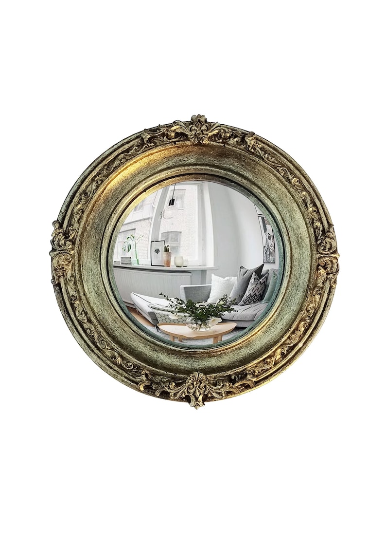May include: A round, antique-style mirror with an ornate, gold-colored frame. The mirror reflects a room with a sofa, plants, and artwork. The frame has detailed floral and scrollwork designs.