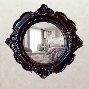 Peut inclure: Un miroir mural rond doté d'un cadre festonné brun foncé orné de détails en relief représentant des motifs floraux et des volutes. La surface en verre réfléchissant montre le reflet intérieur détaillé d'une chambre avec un bureau et un lit.