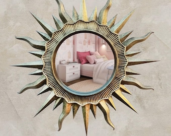 May include: A circular wall mirror featuring a sunburst frame with alternating long and short pointed rays. The frame has a distressed gold and teal finish with a ridged texture surrounding the central glass. The mirror reflects a bedroom interior.