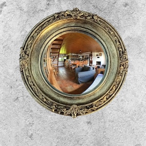 May include: A round, ornate mirror with a gold-colored frame. The frame has intricate detailing and a distressed finish. The mirror reflects a room with a wooden ceiling, staircase, and furniture. The mirror is mounted on a textured, gray wall.