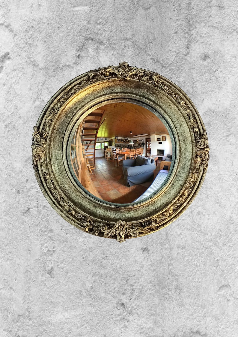 May include: A round, ornate mirror with a gold-colored frame. The frame has intricate detailing and a distressed finish. The mirror reflects a room with a wooden ceiling, staircase, and furniture. The mirror is mounted on a textured, gray wall.