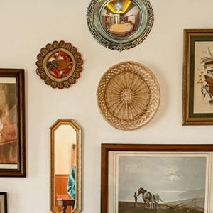 May include: A collection of decorative wall hangings, including a round convex mirror with an ornate gold frame, a woven basket, and framed artwork. The mirror reflects a room. The items are arranged on a white wall.