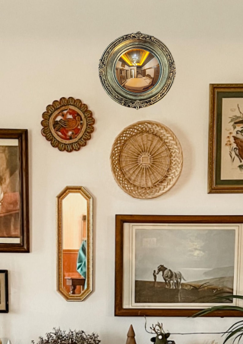 May include: A collection of decorative wall hangings, including a round convex mirror with an ornate gold frame, a woven basket, and framed artwork. The mirror reflects a room. The items are arranged on a white wall.