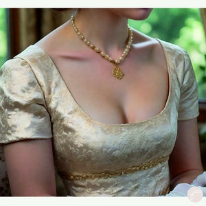 May include: A woman wearing a cream-colored satin dress with a floral pattern. The dress has a scoop neckline and short sleeves. She is wearing a pearl necklace with a gold pendant.