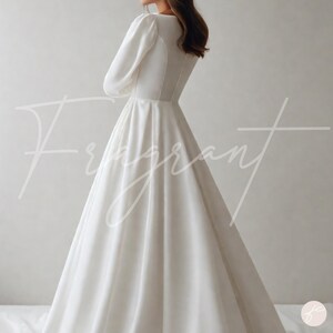 May include: A white, long-sleeved wedding dress with a V-neckline and a flowing skirt. The dress has a fitted bodice and a button detail down the back. The word "Fragrant" is written in cursive across the front of the dress.