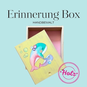 Könnte beinhalten: Eine Erinnerungsbox aus Holz mit einem hellgelben Deckel, der eine handgemalte Illustration eines großen und eines kleinen Wals mit Blumen und Blasen zeigt. Das Innere der Box ist rosa gestrichen. Der Text lautet Erinnerung Box HANDBEMALT.