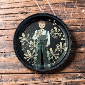 May include: A round, black ceramic plate with a hand-painted design. The plate features a central illustration of a person holding flowers, surrounded by stylized floral motifs. The plate's rim is a darker shade of blue.
