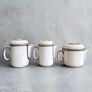 Peut inclure: Trois théières en céramique blanche avec des bordures et des poignées marron foncé sont disposées sur une surface grise. Chaque théière a un couvercle et un bec verseur. Les théières varient légèrement en taille.