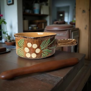 May include: A brown ceramic bowl with a handle, decorated with green leaf and white dot patterns. A wooden rolling pin rests on the table in front of the bowl. The bowl is approximately 10 inches wide.